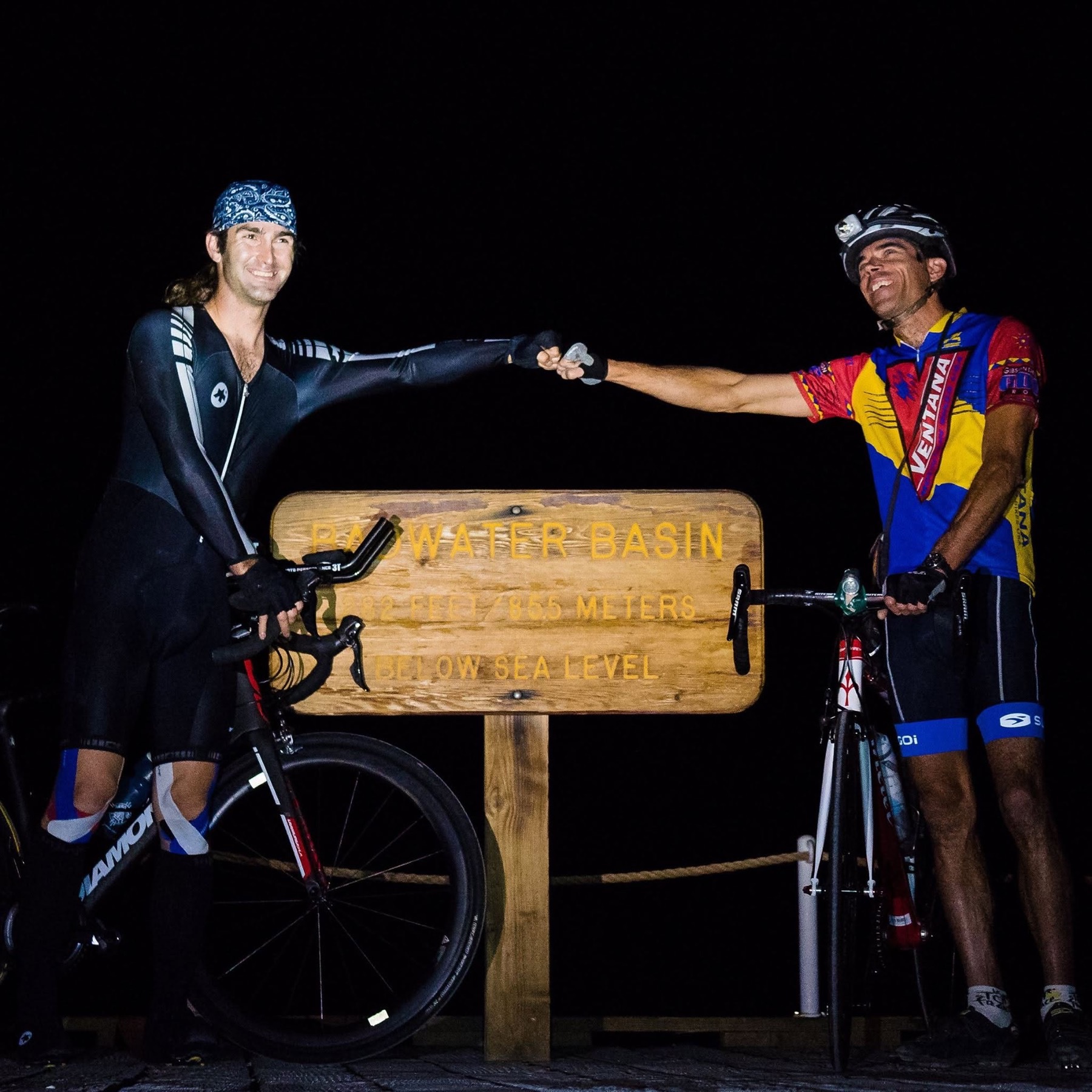 Ryan Tetz and Brent Obinger fist-bump beside their bicycles at the Badwater Basin sign at night