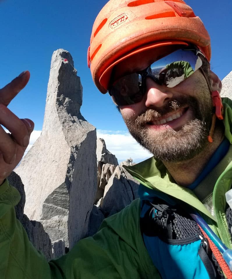 Ryan Tetz smiles in a climbing helmet beside a granite summit block in the California fourteeners