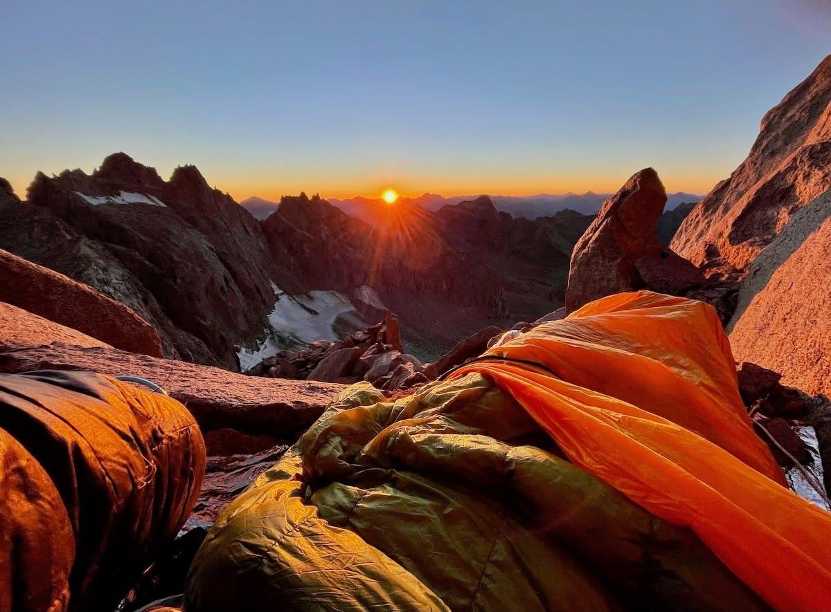 Orange sleeping quilts on a granite bivouac ledge below Mount Sill at sunrise, with North Palisade, Starlight and Thunderbolt beyond