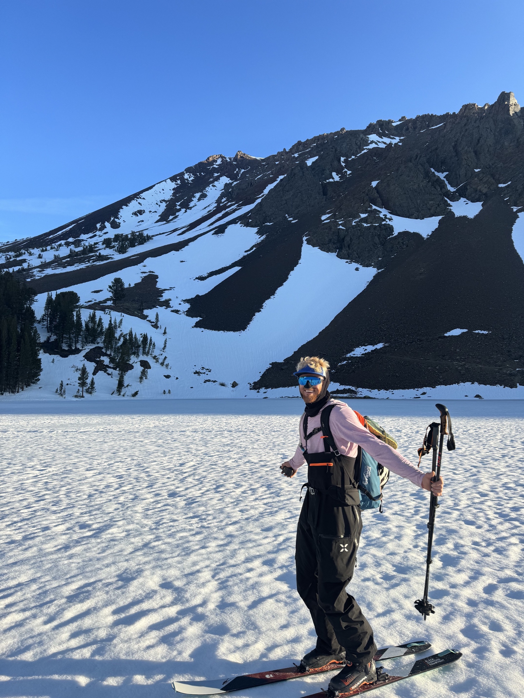 Jake Whisenant smiles on touring skis beneath a snow-streaked mountain ridge on a clear spring morning