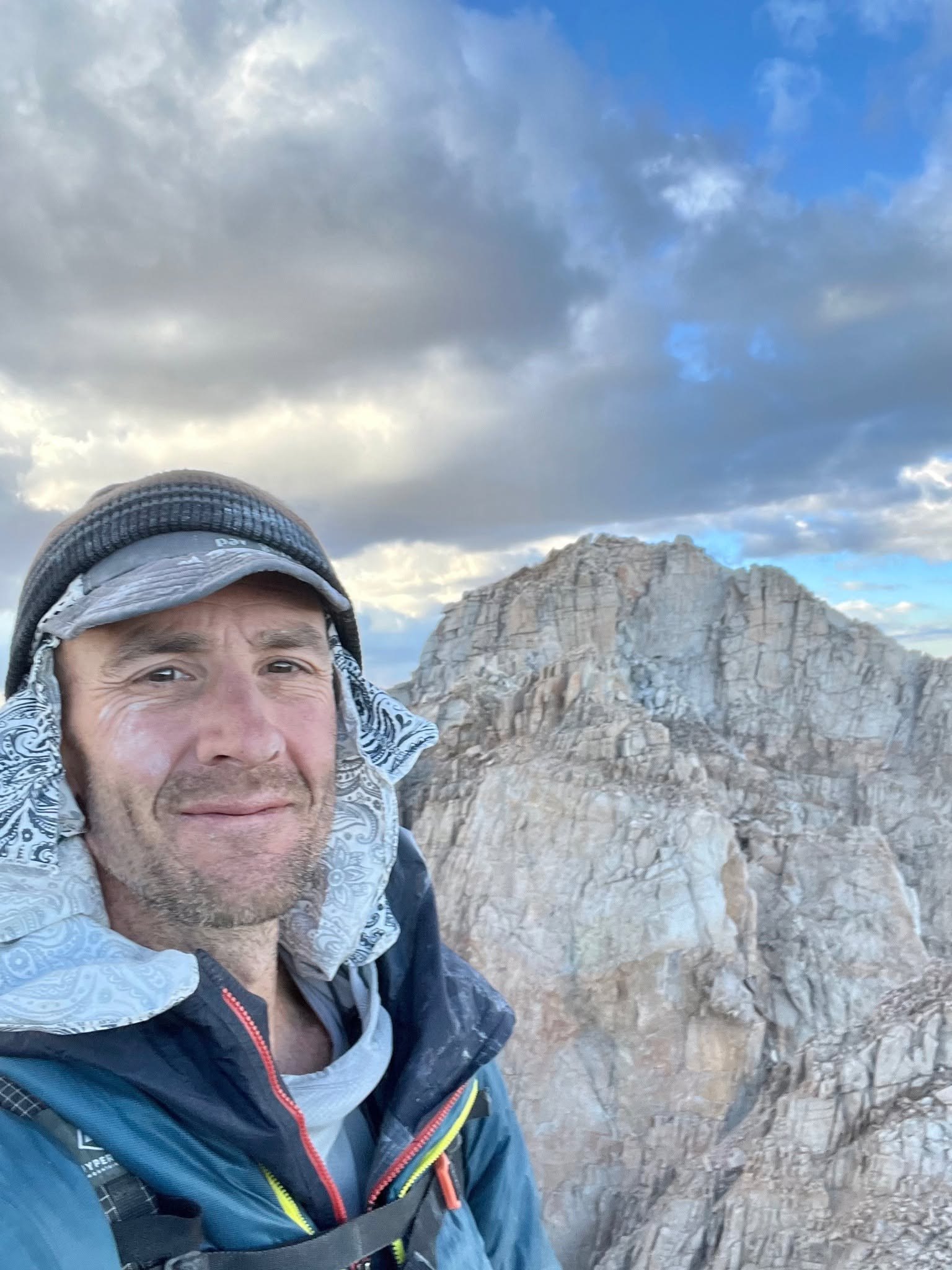 Jason Lakey takes a selfie on a high granite ridge with a blocky summit behind him