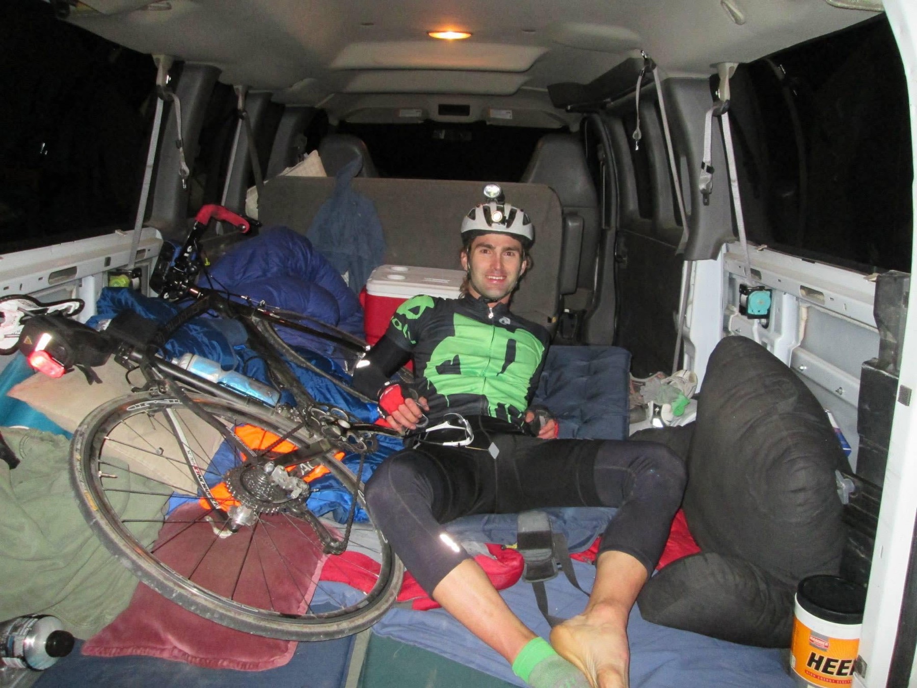 A cyclist rests beside a road bicycle and sleeping system inside the Trans-California support van at night